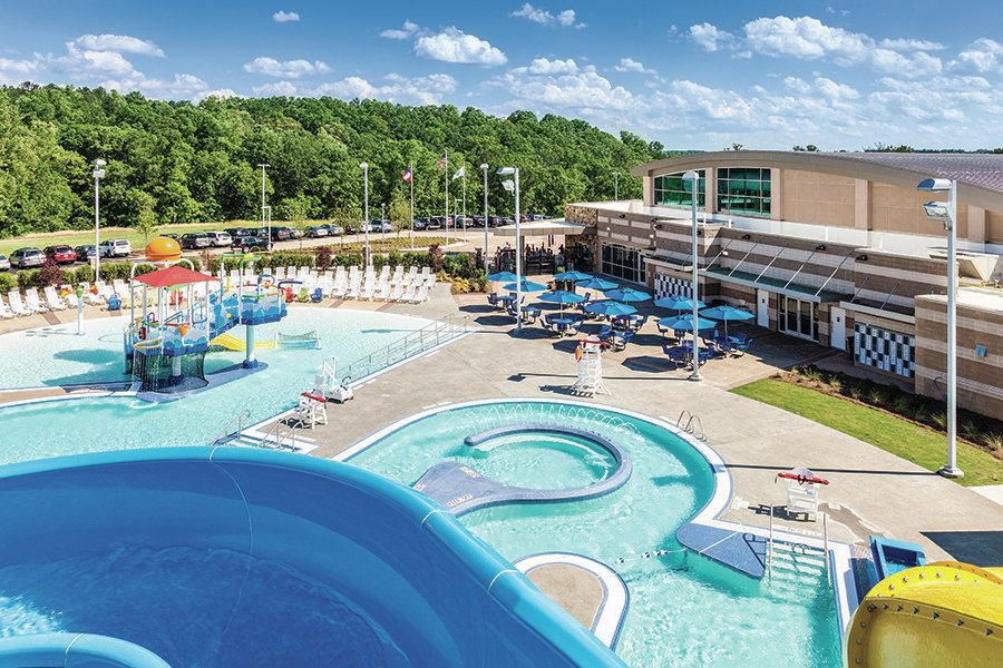 To adhere to the design aesthetic, the lazy river, water sprays, and water slides were placed at the front of the facility. To blend with the setting, natural stone is used at the lobby entry, with wall colors to match. A large window area helps integrate the outdoors with inside, while providing natural day lighting. The curved, wave-like roof form evokes water and helps soften the building so it melds into the tree-lined surroundings.