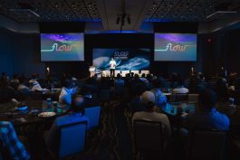 A speaker presents at the SURF PARK SUMMIT to a seated audience in a conference room, featuring a backdrop with colorful "flow" text and ocean imagery.
