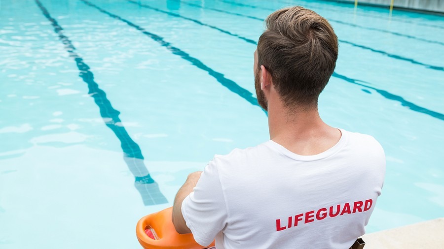 Lifeguard in a white shirt sits beside a turquoise lap pool while holding an orange rescue float.