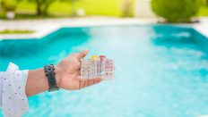 Hand holding a pool water test kit over a bright turquoise swimming pool.